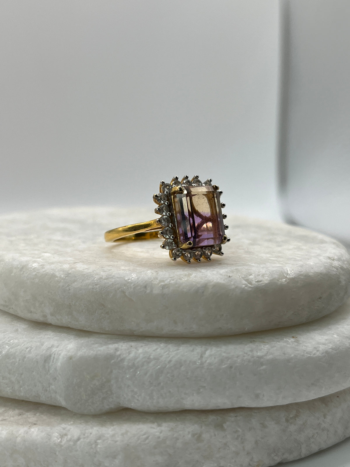 A yellow gold ring featuring a rectangular purple amethyst gemstone surrounded by small clear accent stones, displayed on a white textured ring holder.