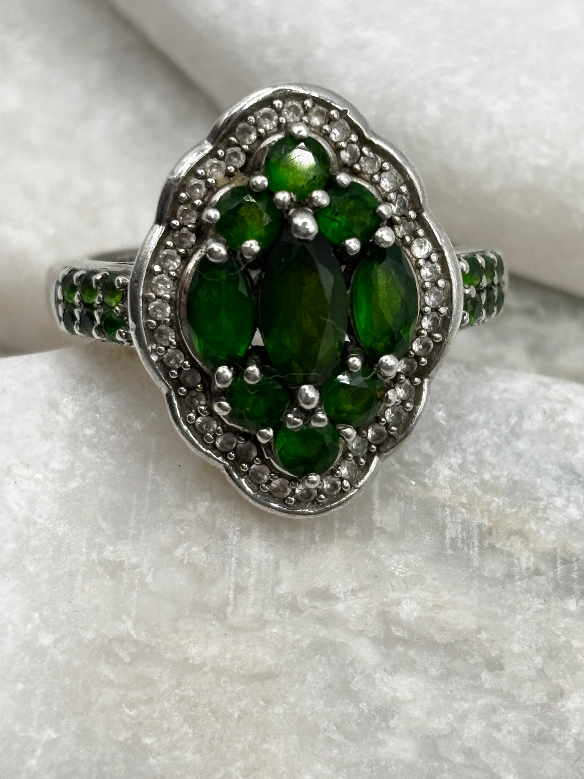 A silver-toned cluster ring featuring a central oval green gemstone surrounded by smaller marquise-cut green stones and a halo of clear stones, displayed on a textured white surface.
