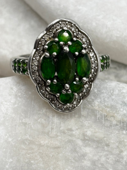 A silver-toned cluster ring featuring a central oval green gemstone surrounded by smaller marquise-cut green stones and a halo of clear stones, displayed on a textured white surface.