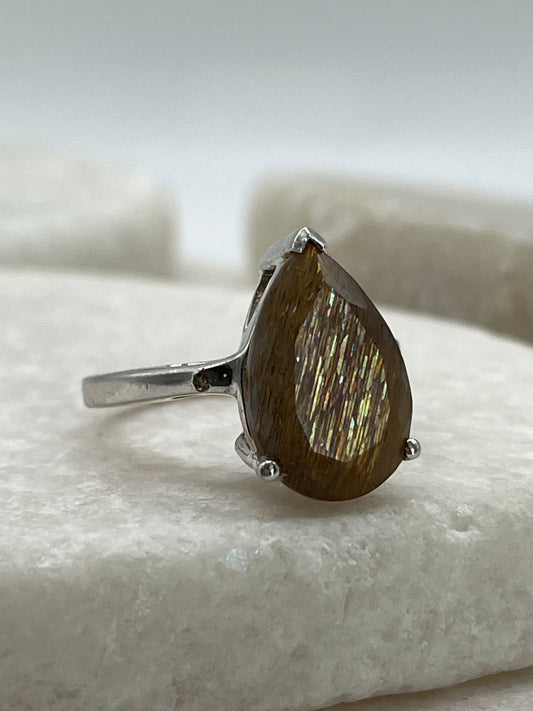 A silver ring featuring a teardrop-shaped golden rutile gemstone with iridescent golden shimmer, displayed on a white textured stone surface.